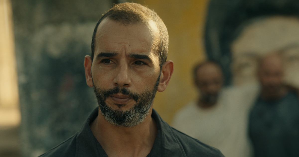 A serious man with a trimmed beard stands outdoors in front of a blurred mural and two other men, with soft natural lighting and a contemplative atmosphere.