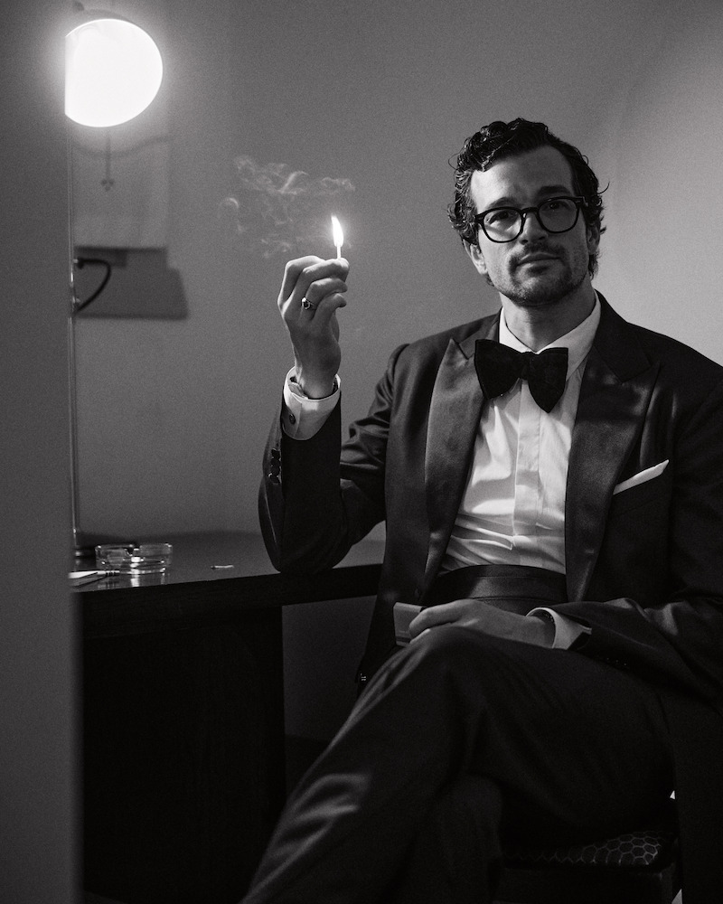Man in a tuxedo sitting at a desk in a dimly lit room, holding a lit match near his face, with a lamp and ashtray on the desk, appearing reflective and calm.