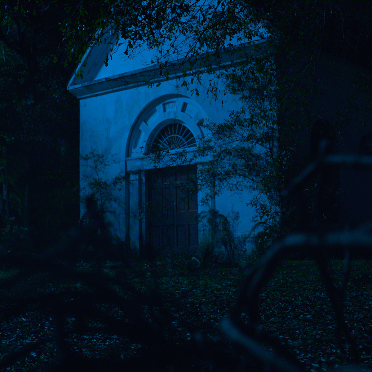 Antigo imóvel branco e de pedra com uma porta de madeira está cercado de árvores à noite, iluminado pelo azul do luar, criando um clima misterioso e assustador.