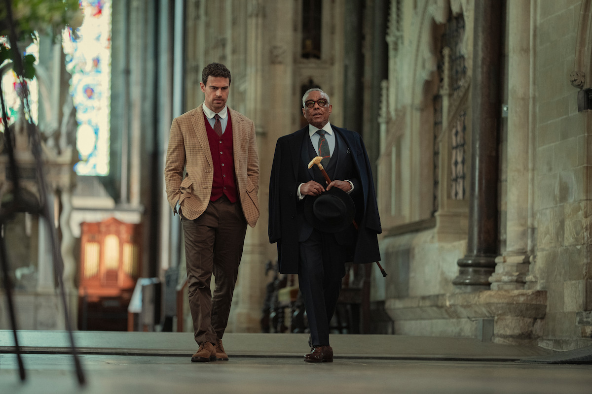 Two well-dressed men walking together inside a grand, historic stone building with high ceilings and stained glass windows, suggesting a church or cathedral setting.