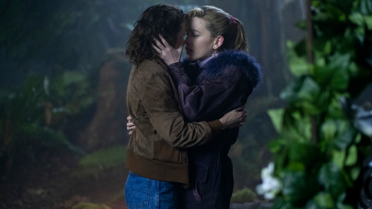 Two women sharing a romantic kiss in a misty, dimly lit forest surrounded by greenery and plants, creating an intimate and emotional atmosphere.