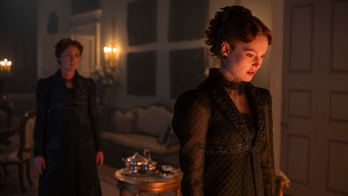 Two women in dark Victorian dresses stand in a dimly lit, elegant room with classical furniture, a tea set on a small table, and candles glowing softly in the background. The atmosphere appears tense and dramatic.