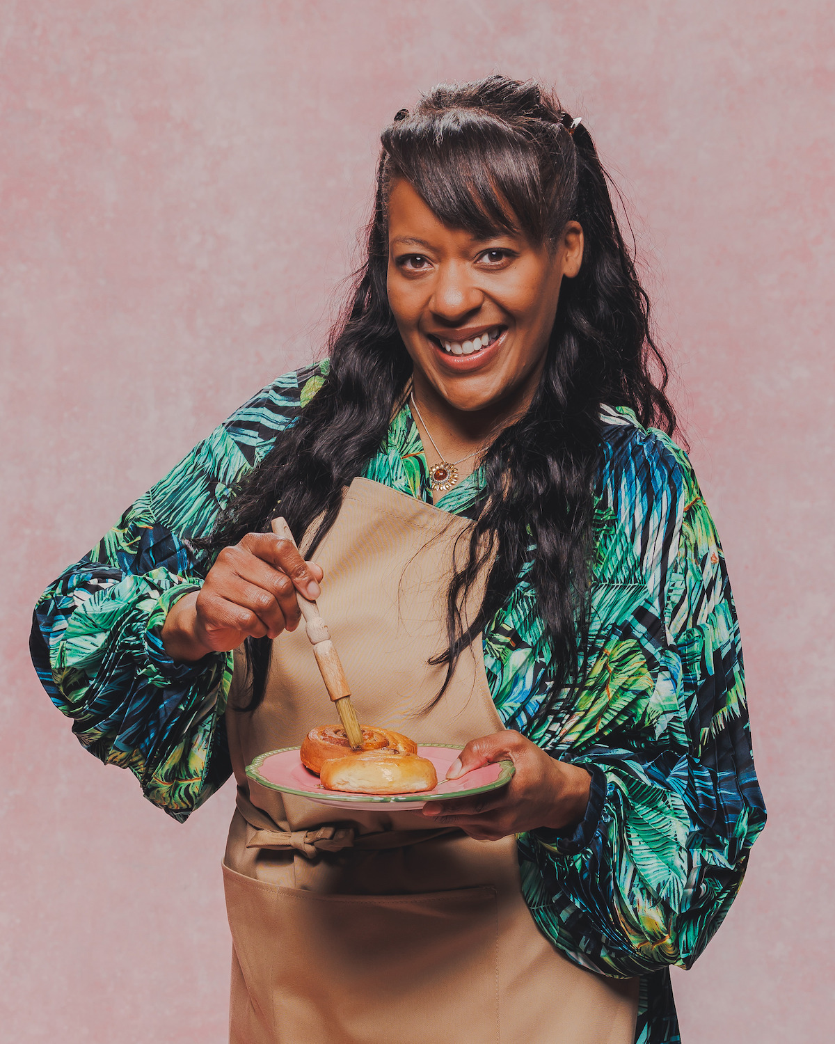 Woman in a tan apron and green patterned shirt smiling while glazing pastries on a plate, standing against a pink textured background in a studio setting.