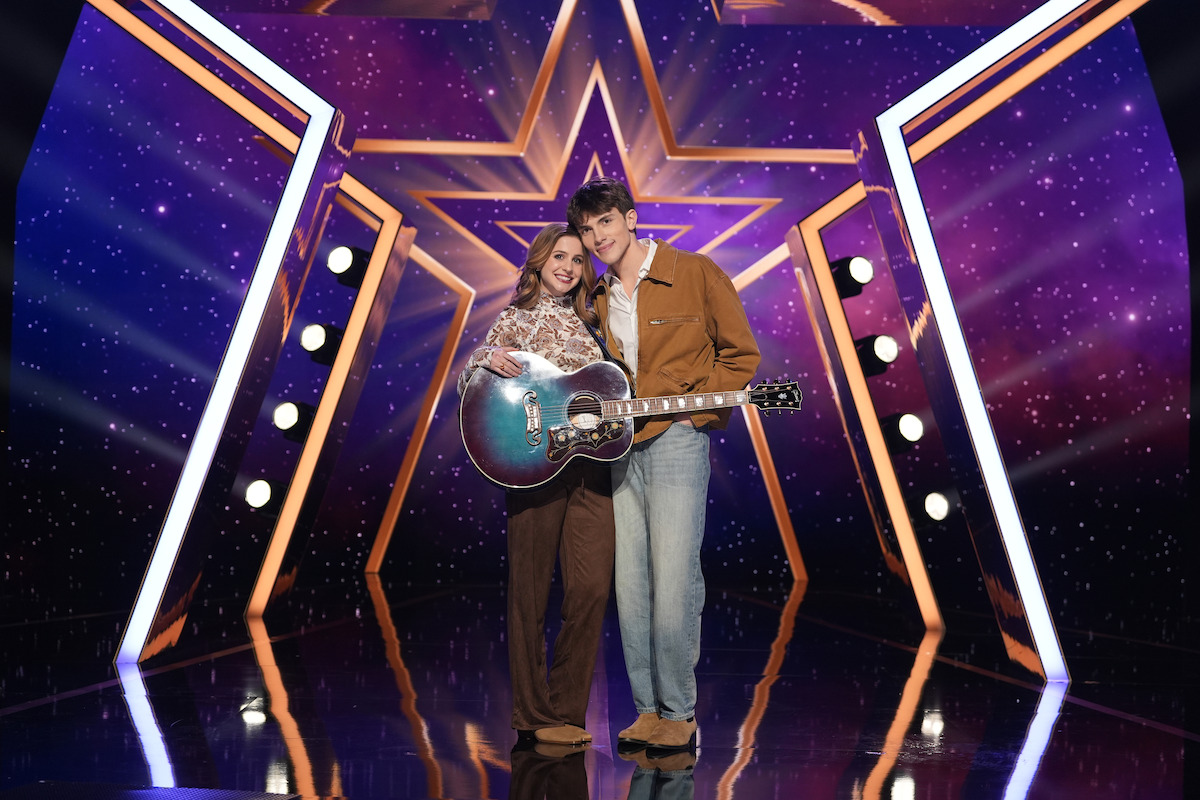 Stuck in September stand together on a brightly lit stage with a large star backdrop. One holds a guitar, both are smiling, and the setting features dramatic lights and a vibrant, colorful environment, suggesting a music or talent show.