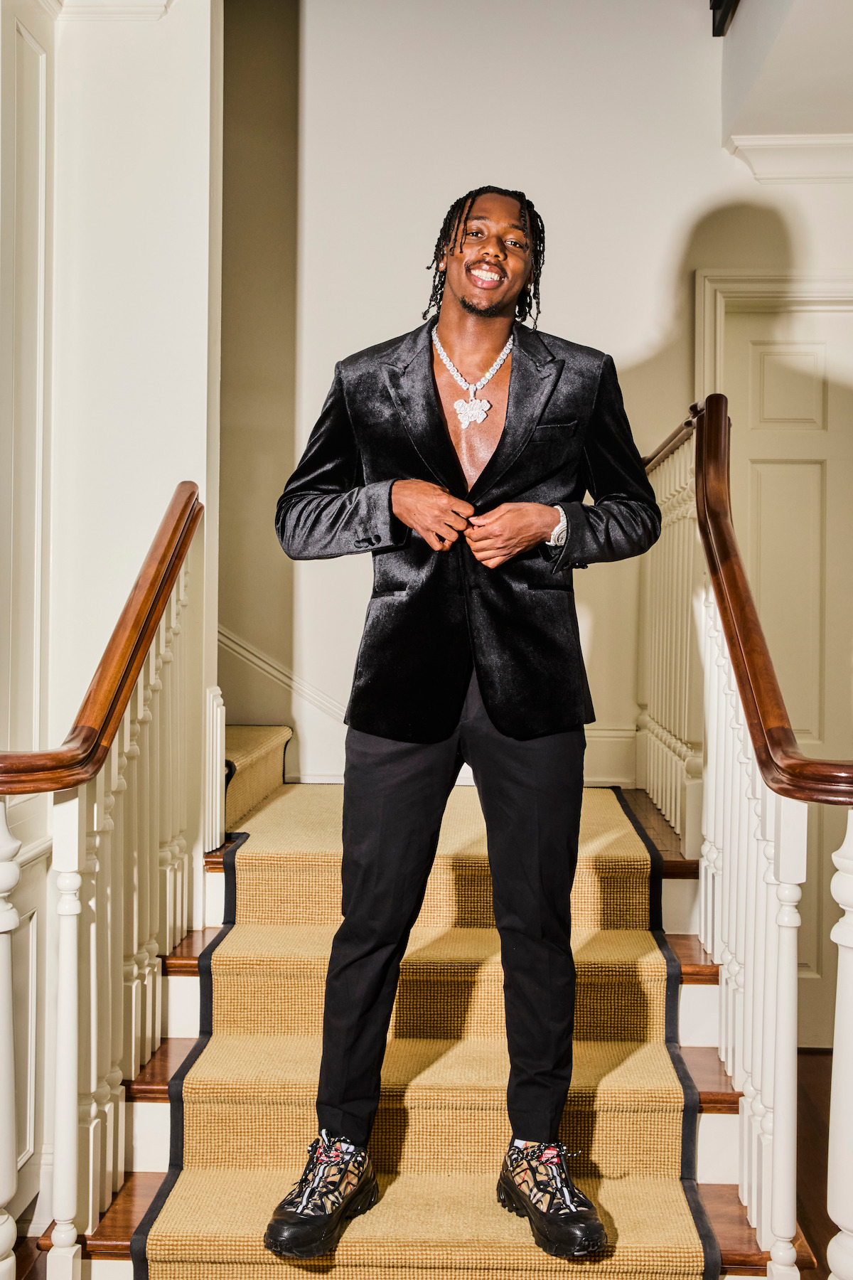 Man in a black velvet blazer, dark pants, and designer sneakers standing confidently on a carpeted staircase in a well-lit, elegant home interior with white walls and wooden railings.