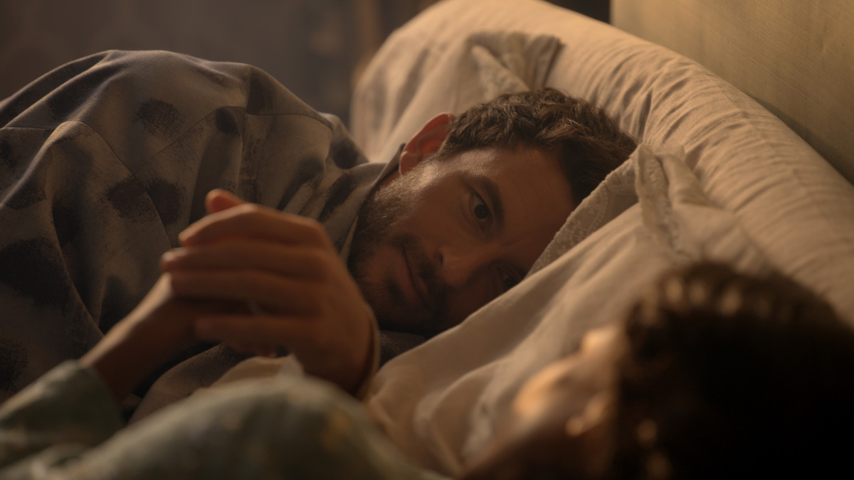 A man and woman lying in bed facing each other in a softly lit bedroom, holding hands, suggesting an intimate and relaxed morning or evening atmosphere.