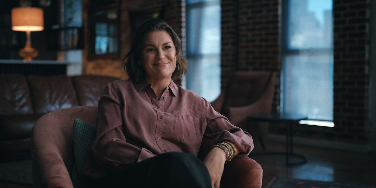 Smiling woman sits in a cozy, modern living room with exposed brick walls, large windows, and soft lighting. She wears a mauve blouse and black pants, with a relaxed, comfortable posture and warm atmosphere around her.
