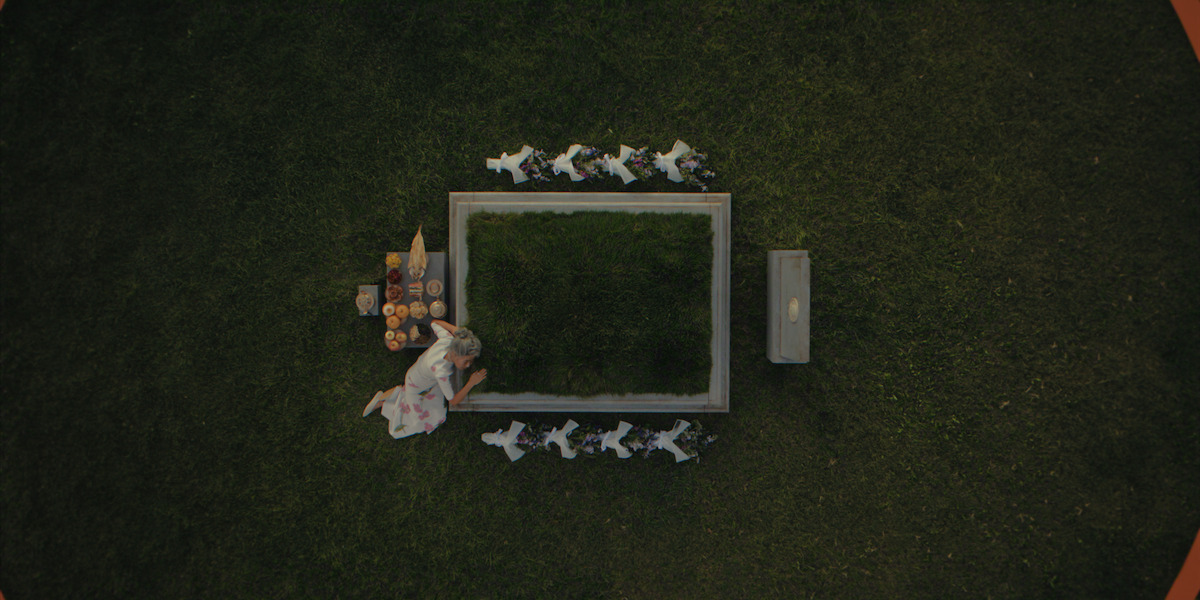A woman in a white dress kneels by a grave in a grassy field, surrounded by small white animal figures and a bench. There are various items placed on the ground near her, giving a ceremonial or memorial atmosphere.
