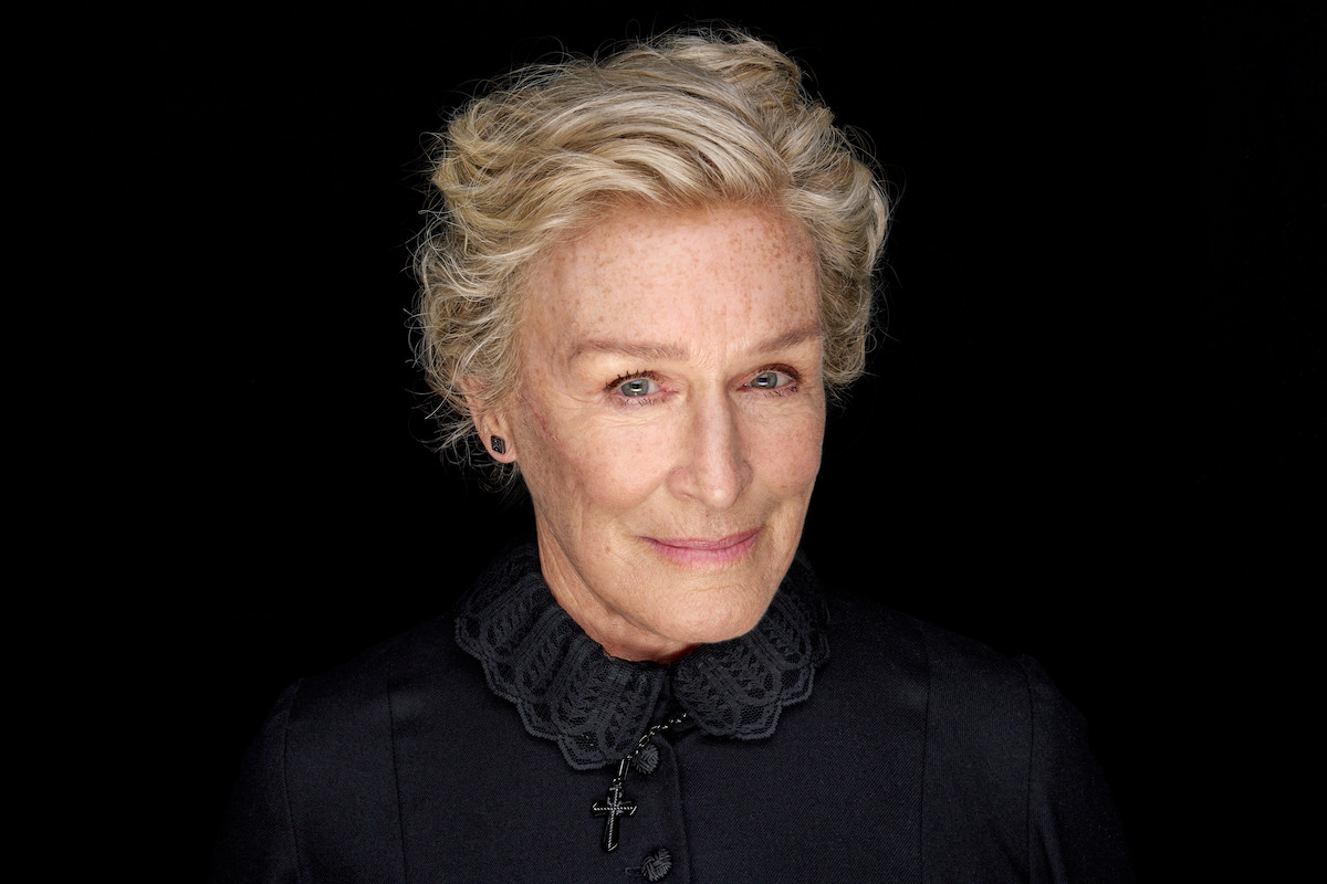 Elderly woman with light hair and a gentle smile, wearing a dark collared garment with a cross necklace, set against a solid black background, creating a calm and dignified atmosphere.