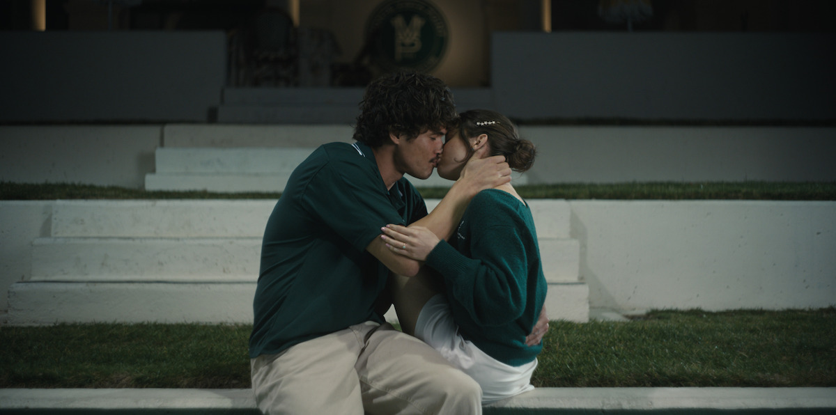 A young couple wearing green and white uniforms sits on outdoor steps, sharing a kiss in front of a lit building at night, with green grass and white stairs surrounding them.