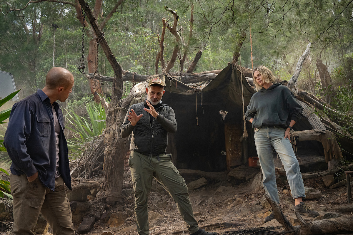 Three people stand and talk in a wooded outdoor setting, in front of a rustic makeshift shelter made of branches and tarps, surrounded by trees and natural vegetation.