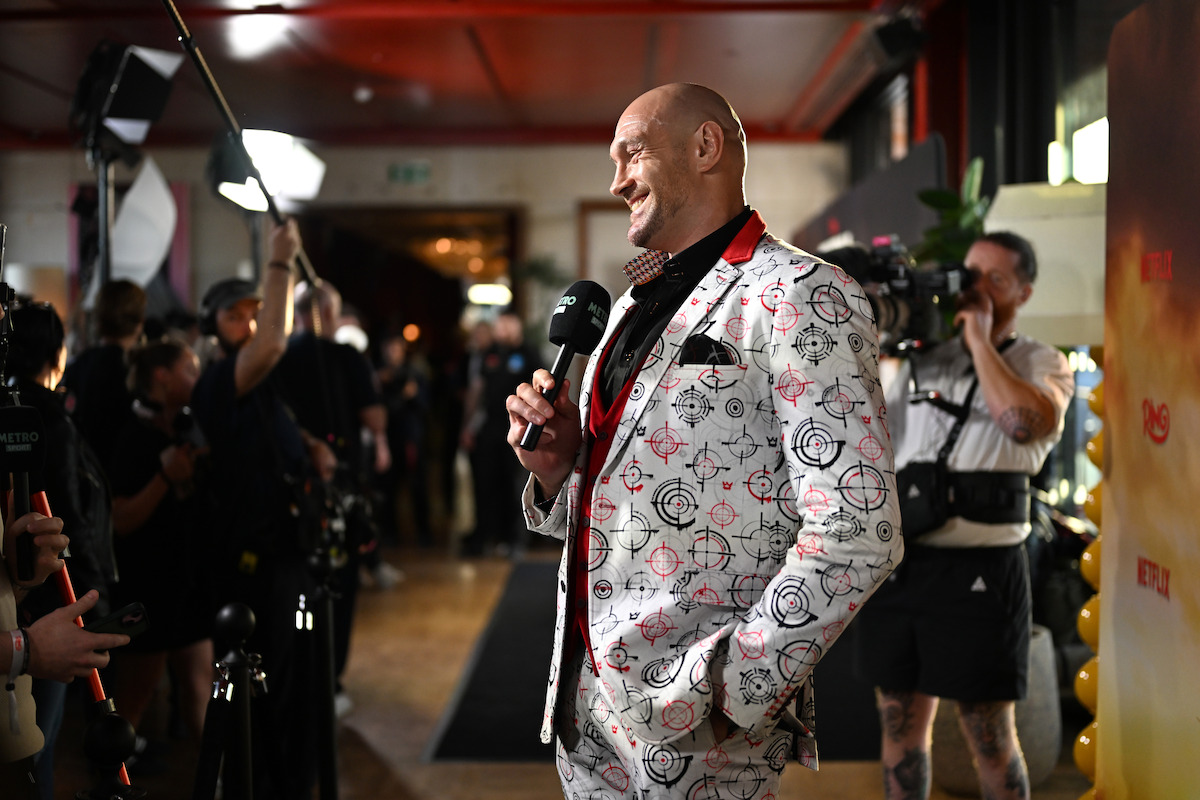 Man in a patterned suit gives an interview on a red carpet, surrounded by photographers, reporters, lighting equipment, and cameras in a lively indoor event setting.