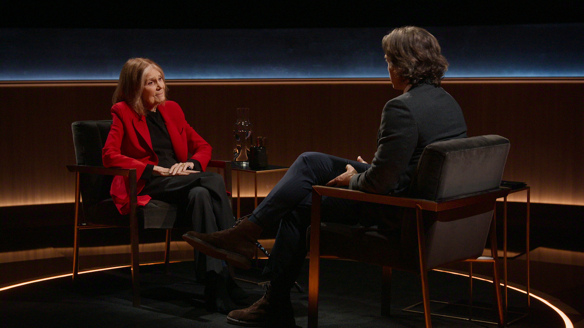 Two people sit facing each other in armchairs on a dimly lit stage, having a conversation. The woman wears a red blazer, and a table with glasses is between them. The environment is modern and intimate, suggesting a formal interview.