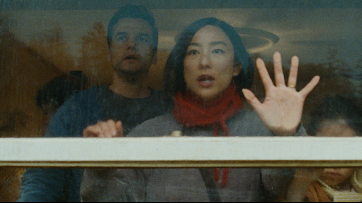 Three people stand indoors, looking out a window with concerned expressions. A woman with a red scarf has her hand raised on the glass, with a man and child beside her. The environment looks tense and reflective, with muted lighting.