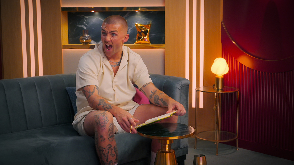 A person with tattoos sits on a modern gray couch in a stylish, warmly-lit room, holding a tablet and reacting with surprise or excitement. Decorative lighting and shelves with art pieces are visible in the background.