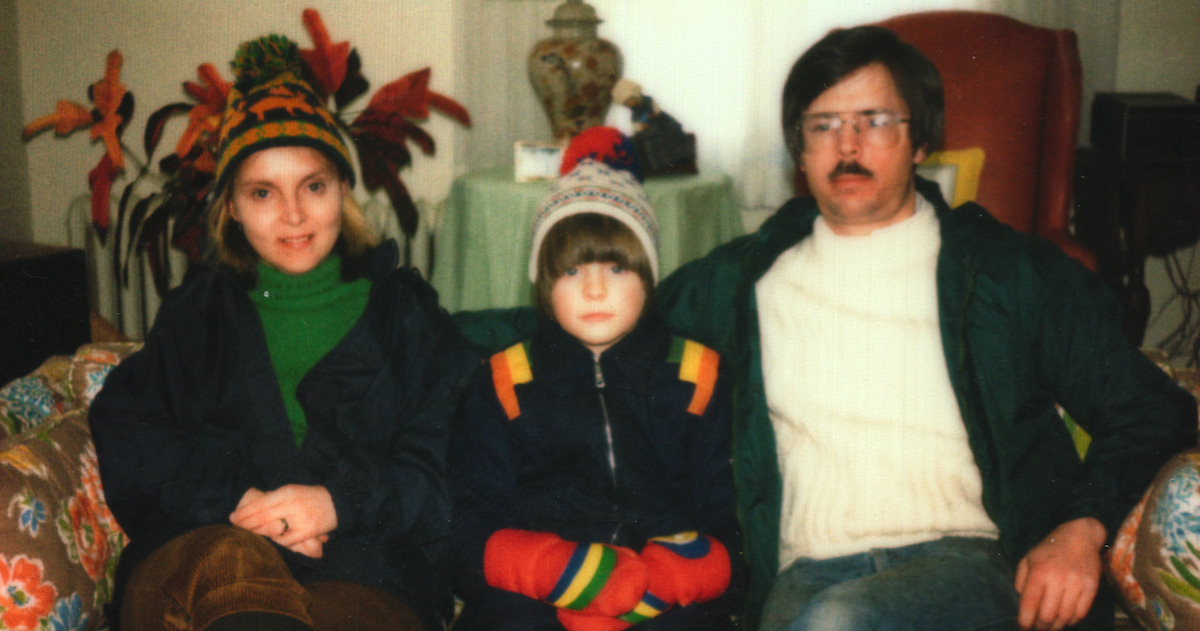 Three people in winter clothing sit on a patterned couch indoors, with houseplants and a lamp on a table in the background, appearing relaxed in a cozy living room setting.