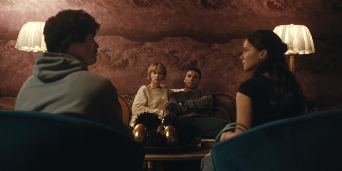 Four people sit in a warmly lit living room with two lamps, plush chairs, and decorative eggs on the table, engaged in an intense conversation against a textured mauve wall.
