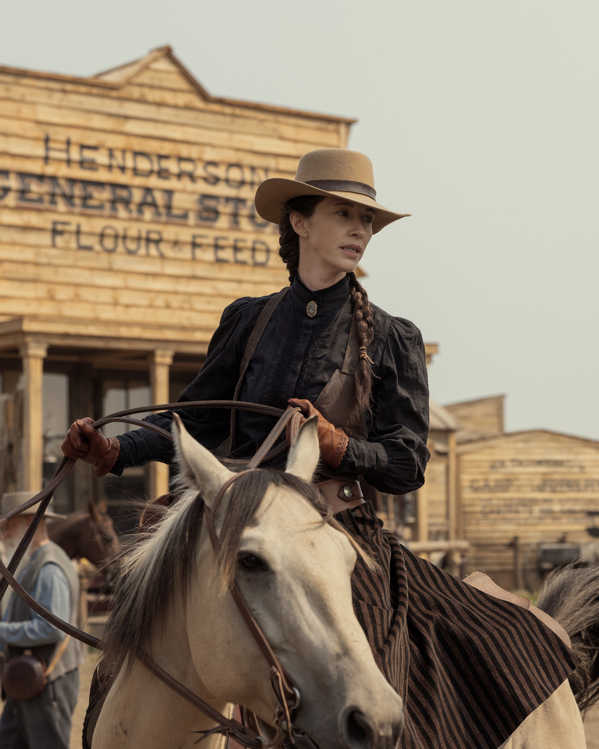 Woman in old-fashioned Western clothing riding a horse in front of a wooden general store with "Henderson General Store Flour Feed" sign, set in a dusty, historical frontier town.