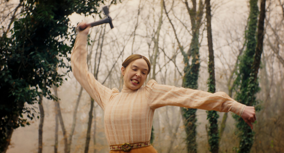Young woman in vintage dress fiercely swinging a hammer in a wooded, misty forest setting, surrounded by tall, leafless trees and ivy-covered trunks, with determined expression and dynamic movement.