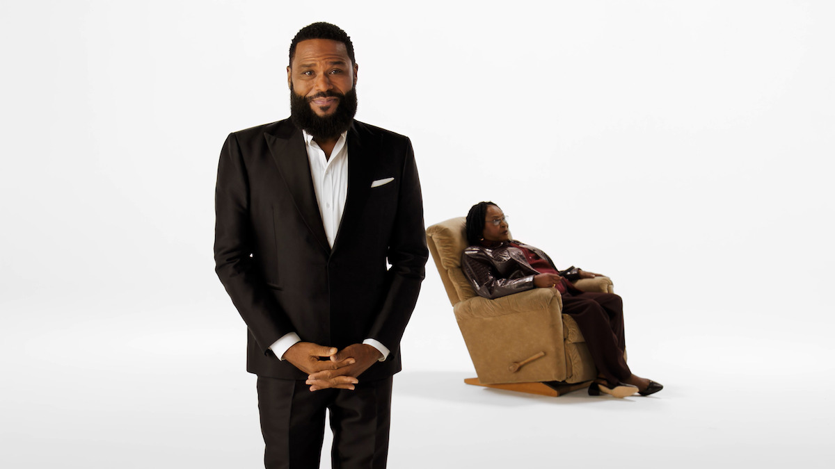 Anthony Anderson, wearing a black suit, stands confidently in the foreground of a white studio, while his mother, Doris Hancox, sits in a recliner in the background, creating a contrast in posture and concentration.