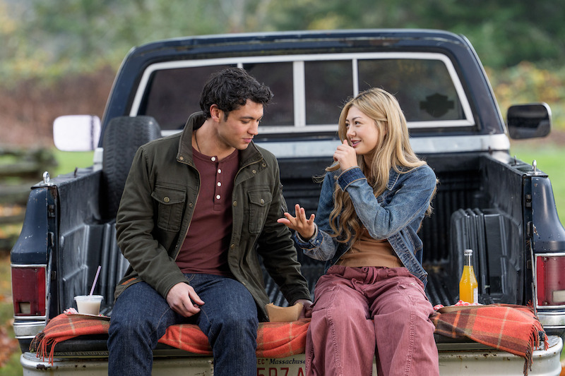 Man and woman sitting on tailgate of a pickup truck in a grassy outdoor setting, talking and eating together, with drinks and food on a blanket, autumn trees in the background.