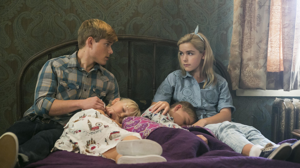 Two older children sit on a bed in a dimly lit room comforting two younger children who are asleep with their heads in their laps, suggesting a caring, protective atmosphere. The room has vintage wallpaper and curtains.