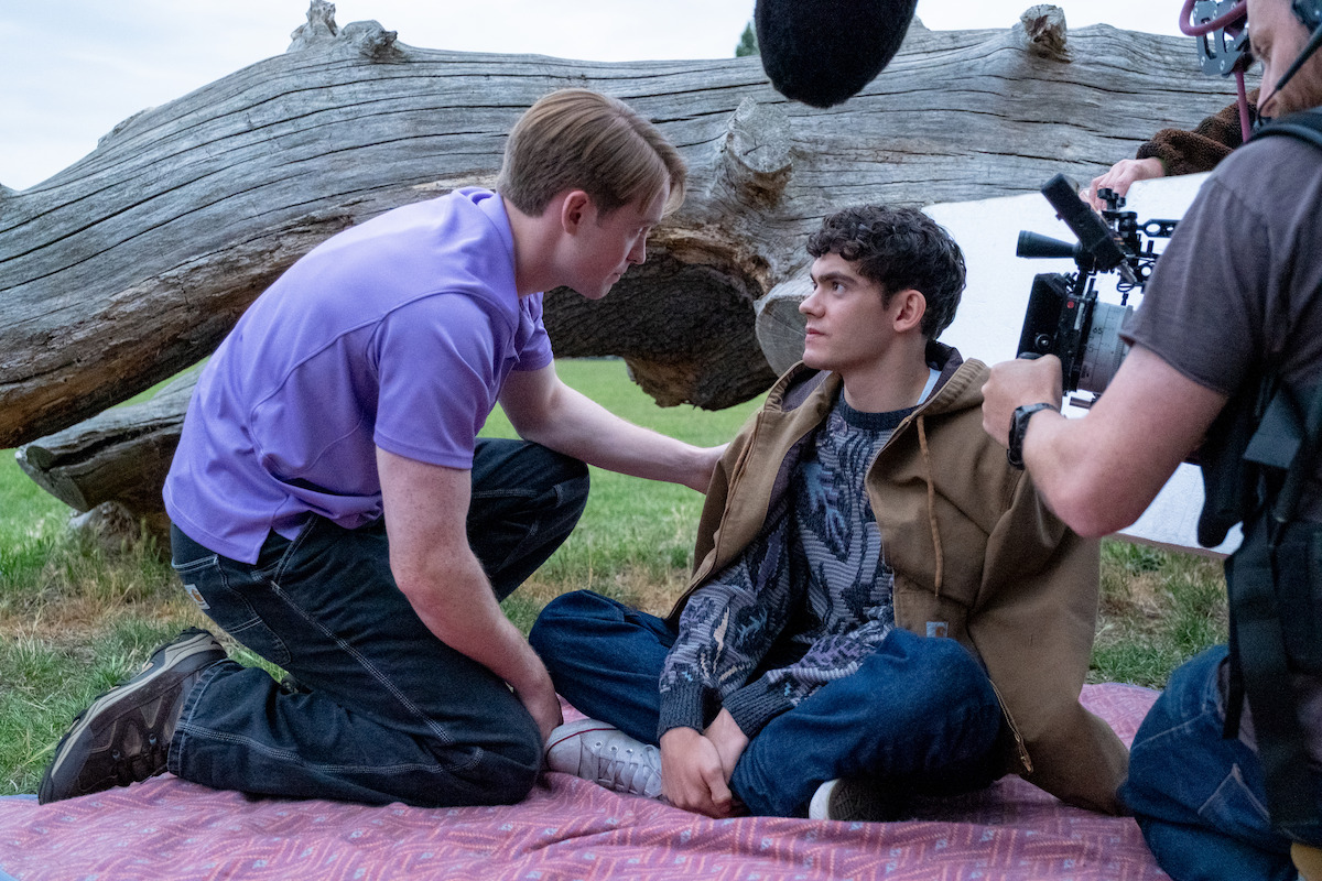 Two young men sit on a blanket outdoors near a fallen tree, one comforting the other, surrounded by filming equipment and crew in a natural grassy setting.