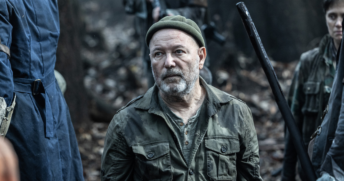 An older man wearing army clothes stands in a battlefield with a forlorn expression.