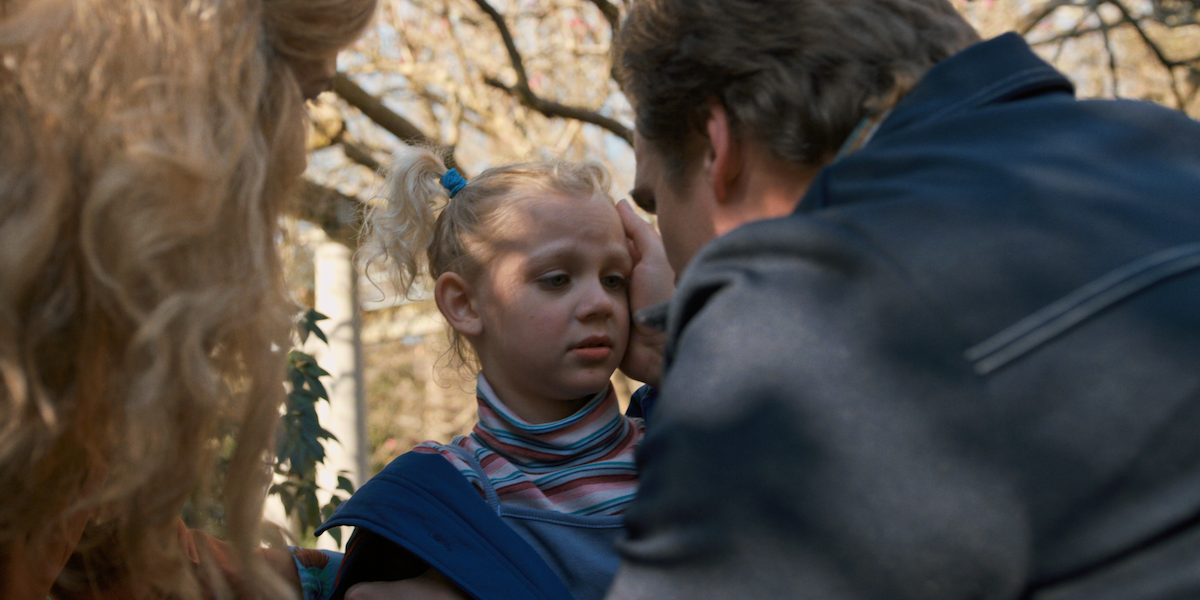 A concerned adult gently comforts a young girl with blonde pigtails outdoors.