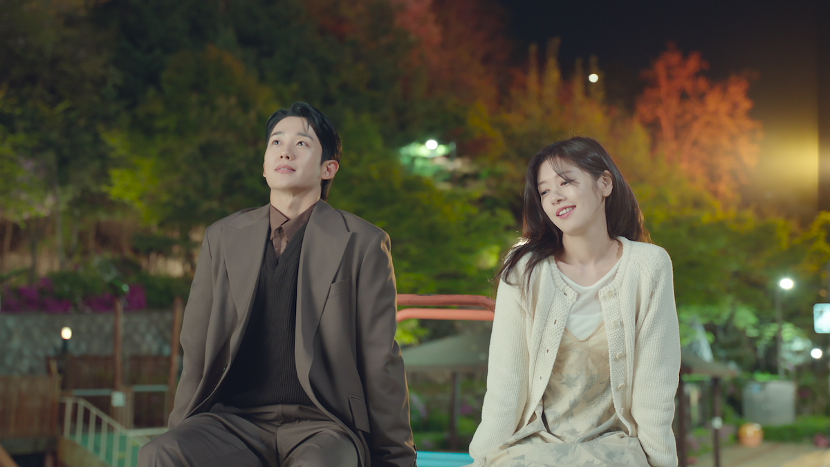 Jung Hae-in as Choi Seung-hyo, wearing a brown suit, sits outside on a bench next to Jung So-min as Bae Seok-ryu, who wears a cream-colored floral dress and cream-colored cardigan in an image from the series ‘Love Next Door.’