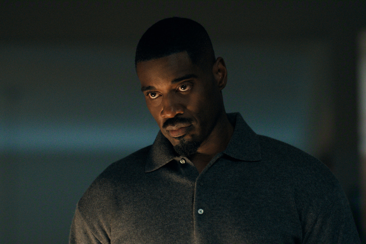 Serious man in a grey collared shirt standing indoors under moody, low lighting, with a neutral background and a focused expression.