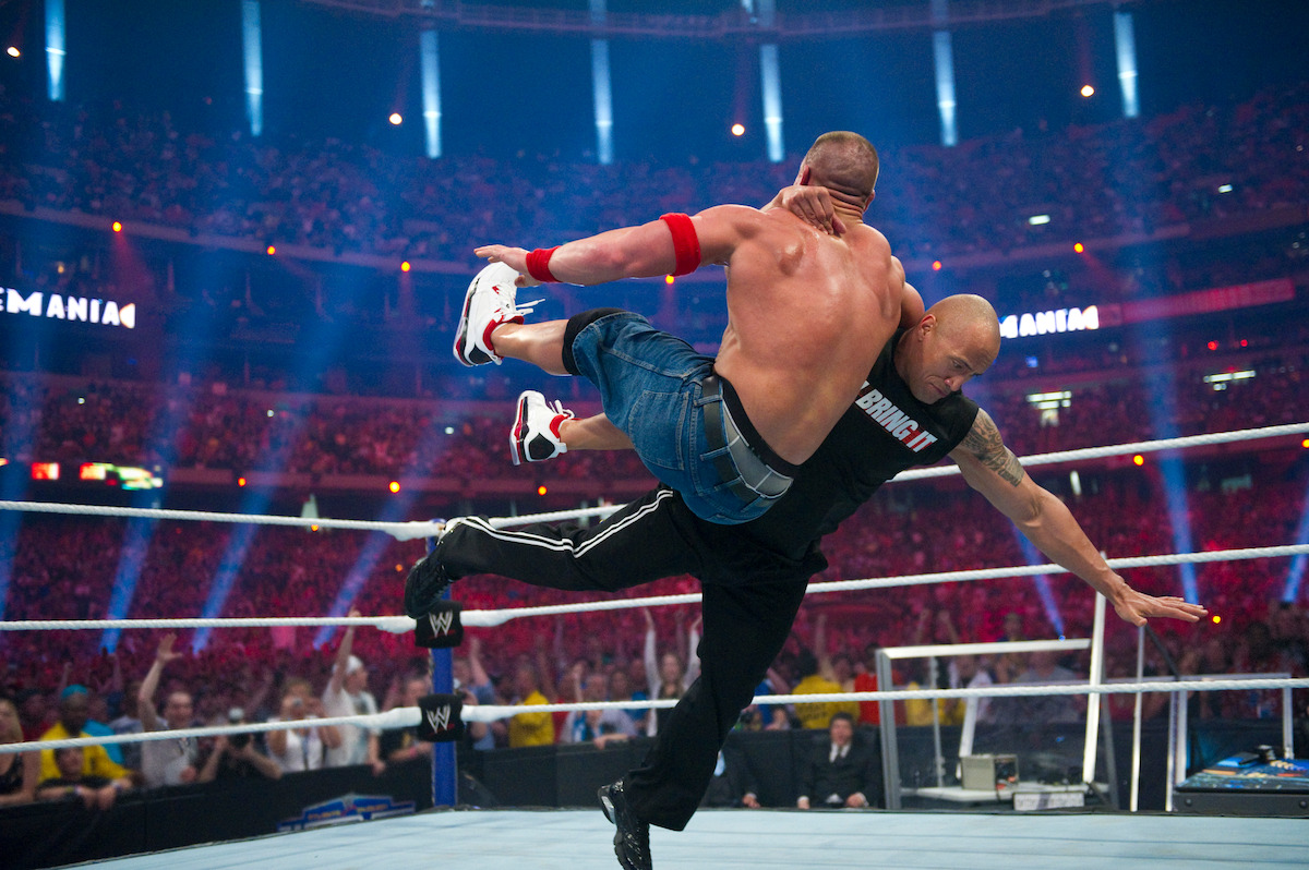 Two muscular wrestlers are engaged in a dramatic match inside a brightly lit wrestling ring, surrounded by a large cheering stadium crowd, capturing the intense action and excitement of the event.