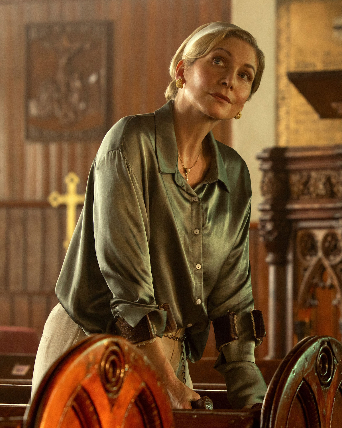 Elizabeth Mitchell as Carla Limbrey blonde woman in a satin blouse and handcuffs stands in an ornate wooden church, with religious symbols and carvings visible, looking upward pensively.