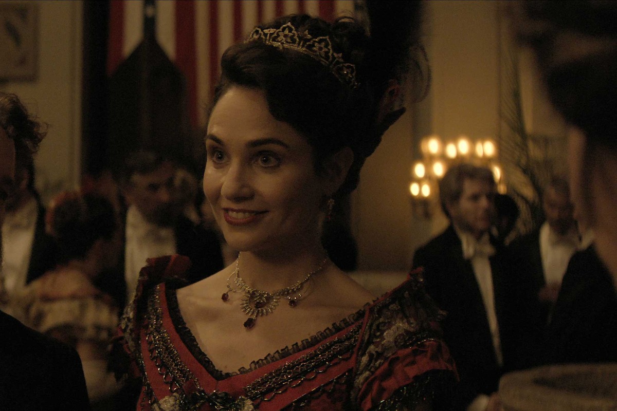 Woman in ornate dress and jewelry smiles at a formal ball; elegant 19th-century setting, warm lighting, guests in evening attire, festive, lively atmosphere.
