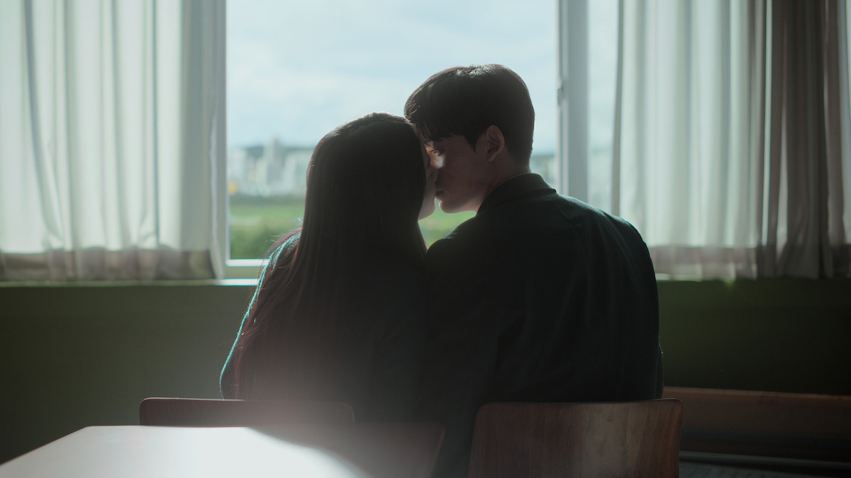 Two people sit closely together at a desk in a softly lit room, facing a large window with sheer curtains, sharing an intimate moment as daylight shines in.