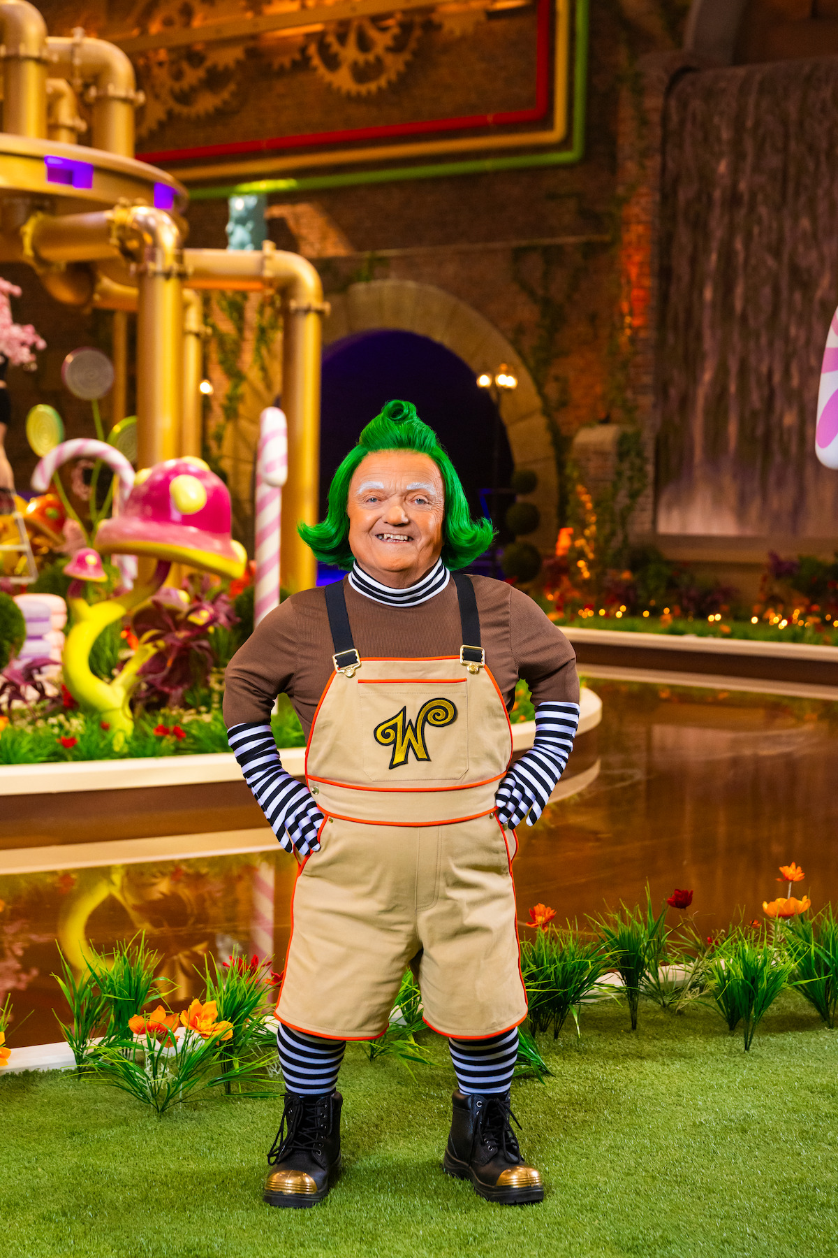 Person dressed as a classic Oompa Loompa with green hair, striped shirt, and khaki overalls stands in a whimsical, colorful chocolate factory set with candy decorations, pipes, and a waterfall in the background.