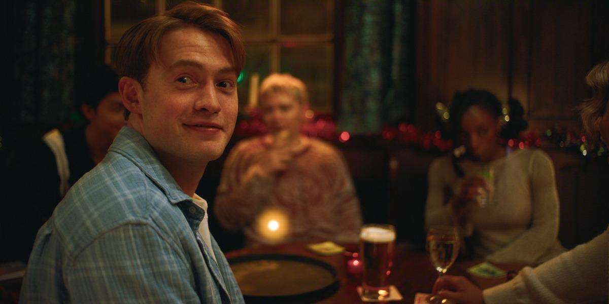 Young man in a plaid shirt smiles while sitting at a cozy pub table with friends. Warm lighting, drinks, and festive decorations create a relaxed, social atmosphere.