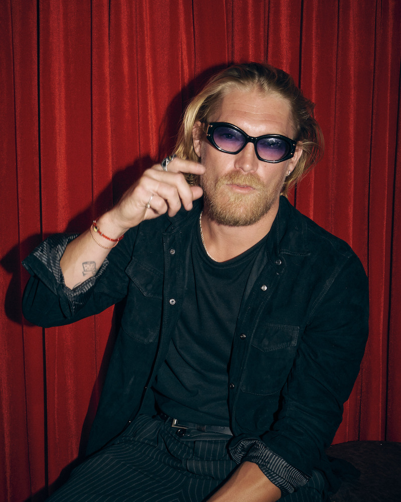 A man with long blond hair, beard, and sunglasses sits in front of red curtains, wearing a black shirt and striped pants, holding his hand near his face in a casual indoor setting.