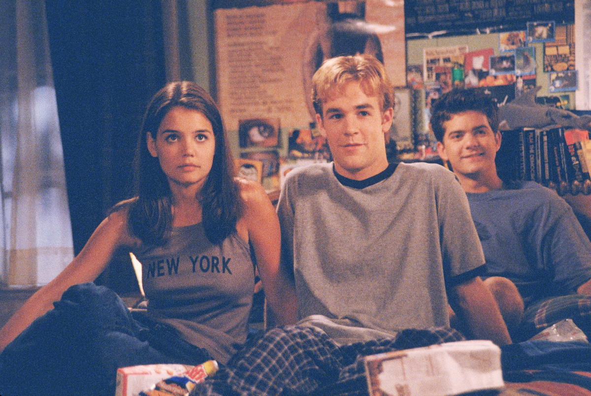 Three teenagers sit closely together in a cozy bedroom with posters and photos on the wall behind them, relaxed and casually dressed, surrounded by personal belongings and snacks, suggesting a friendly and informal setting.