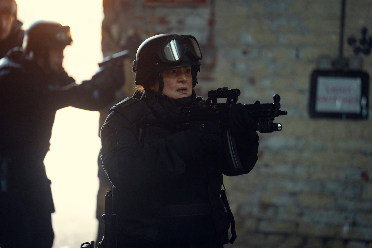 Armed SWAT officers in tactical gear move through a dim, industrial setting with a tense, focused atmosphere, brick wall and hazy light in the background suggesting a high-stakes operation.