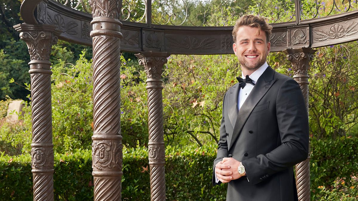 Man in a black tuxedo smiling outdoors, standing near ornate pillars and greenery, with an elegant garden gazebo structure in the background.