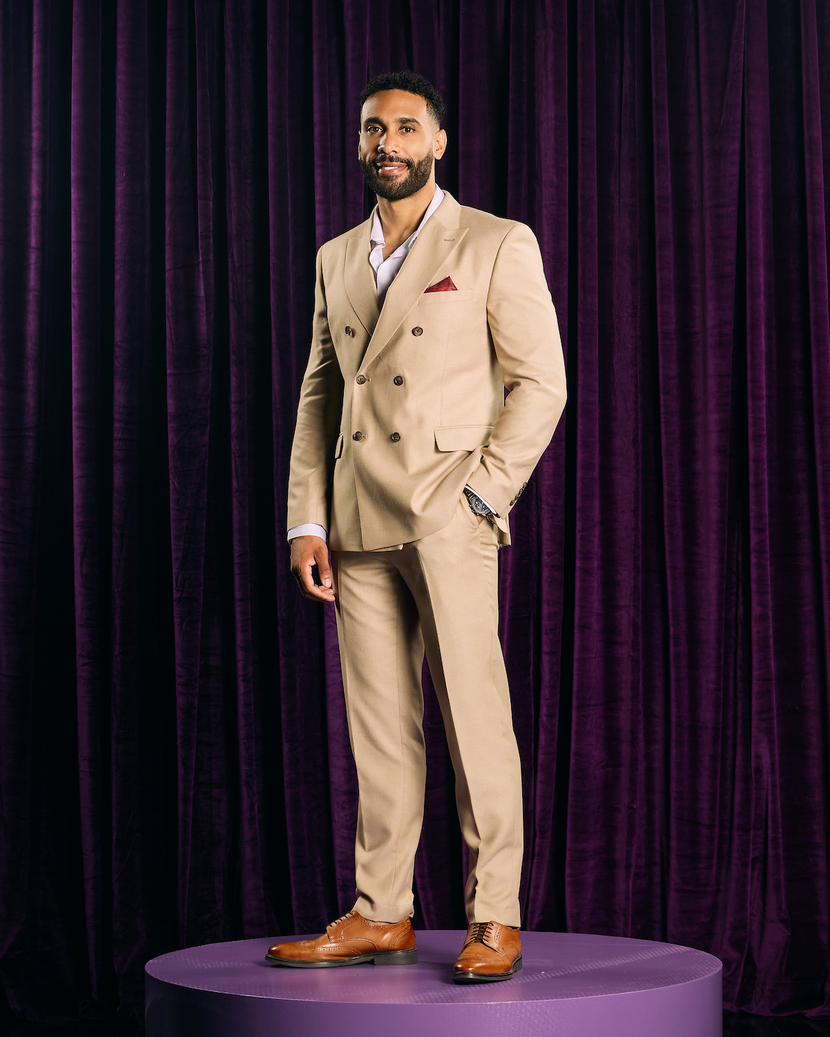 Man in a beige suit with a red pocket square stands confidently on a purple circular platform, posed against a background of rich, dark purple velvet curtains in a studio setting.