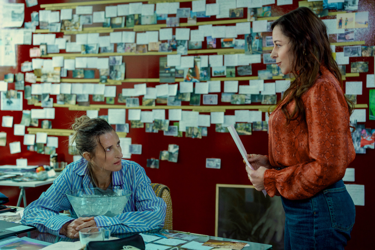 Two women in an office with one seated at a cluttered desk and the other standing, talking. The wall behind them is covered with pinned notes and photographs, suggesting an investigation or research setting.