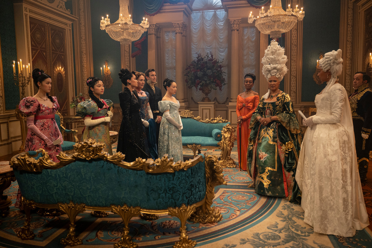 A group of people in elaborate, colorful Regency-style costumes stand in a grand, ornate room with chandeliers, gold-trimmed blue sofas, and rich décor, suggesting a formal gathering or royal event.