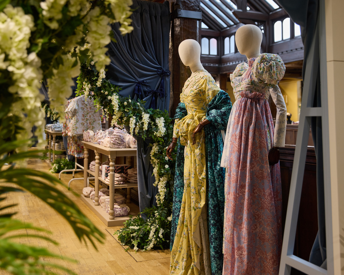 Two mannequins dressed in vintage floral gowns stand in a well-lit, elegant indoor shop decorated with greenery, flowers, and draped fabrics; folded fabric is stacked on tables in the background.