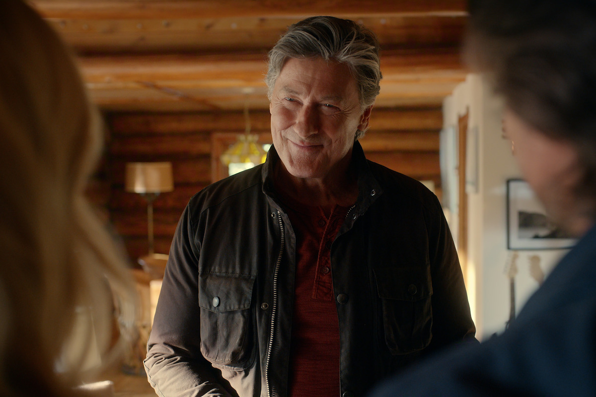 Older man smiling in a warmly lit log cabin, wearing a dark jacket over a red shirt, with two people in the foreground, cozy rustic interior with wooden walls and a lamp in the background.