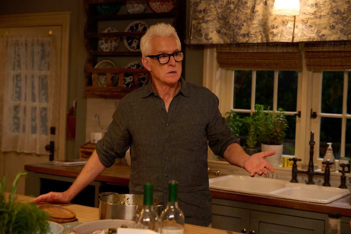 John Slattery, who plays John, stands in a cozy, warmly lit kitchen, gesturing with one hand. The kitchen counter has potted plants, dishes, and bottles, with a window in the background.
