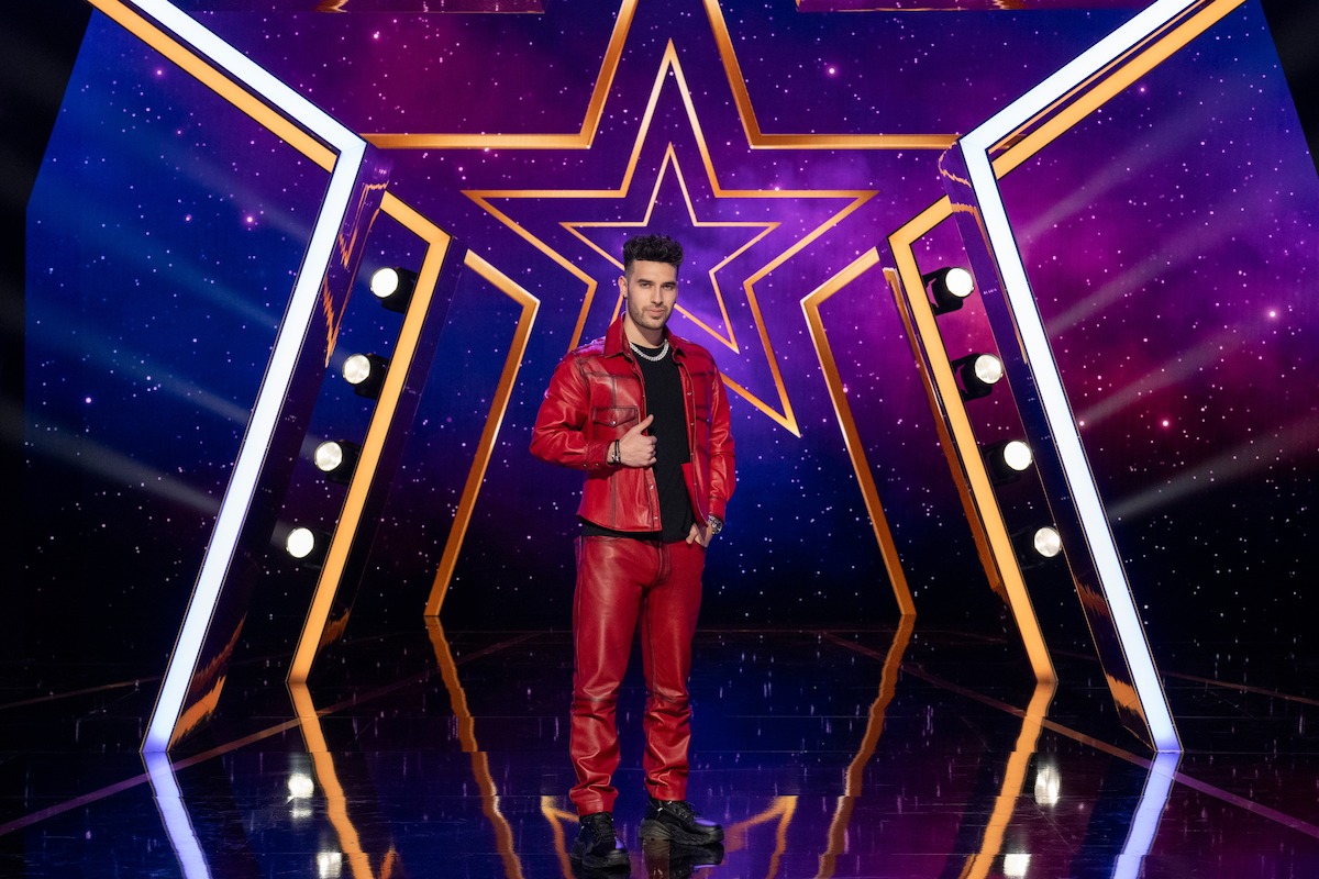 TJ Salta con un traje de cuero rojo posando con confianza en un escenario brillante con luces de neón en forma de estrella y un fondo morado con temática de galaxias, enmarcado por focos brillantes dispuestos en ambos lados. TJ Salta con un traje de cuero rojo posando con confianza en un escenario brillante con luces de neón en forma de estrella y un fondo morado con temática de galaxias, enmarcado por focos brillantes dispuestos en ambos lados.