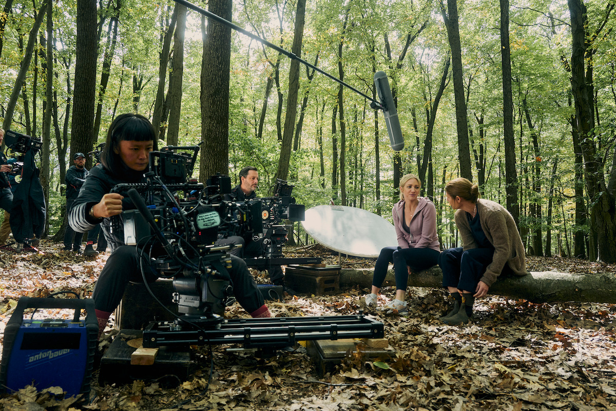 Film crew shoots a scene in a forest, with actors sitting on a log talking, surrounded by cameras, crew, equipment, and a large microphone, creating a focused, behind-the-scenes filmmaking atmosphere.