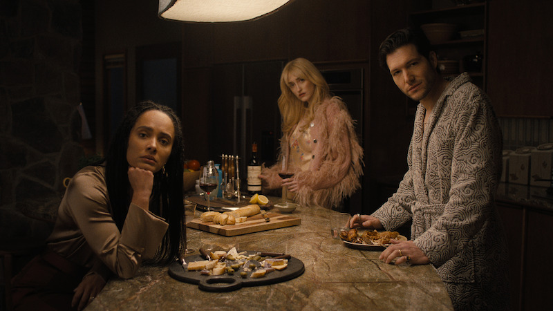 Three people in stylish clothing stand around a modern kitchen island at night, with drinks, food, and snacks on the counter, illuminated by warm overhead lighting, creating a cozy yet tense atmosphere.